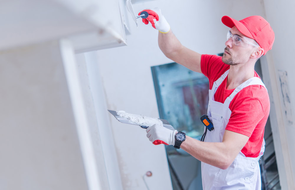 A worker wearing a red cap and shirt, white overalls, and safety goggles is plastering a wall in a room likely under renovation. With a trowel in one hand, he smooths the surface with the other. This meticulous interior painting occurs in Suffolk County.