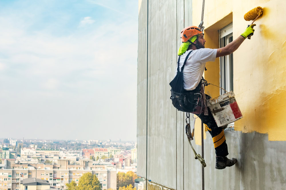 A worker in safety gear is suspended by ropes, expertly handling a roller for exterior painting on a high-rise. Below lies the sprawling cityscape of Suffolk County, showcasing various buildings under a vibrant blue sky.