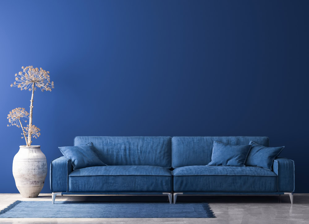 A minimalist living room featuring a deep blue sofa with cushions on a matching blue rug. A large white vase with dried plants stands on the left. The bold blue wall, enhanced by expert interior painting from Nassau County, creates a monochromatic and serene atmosphere.