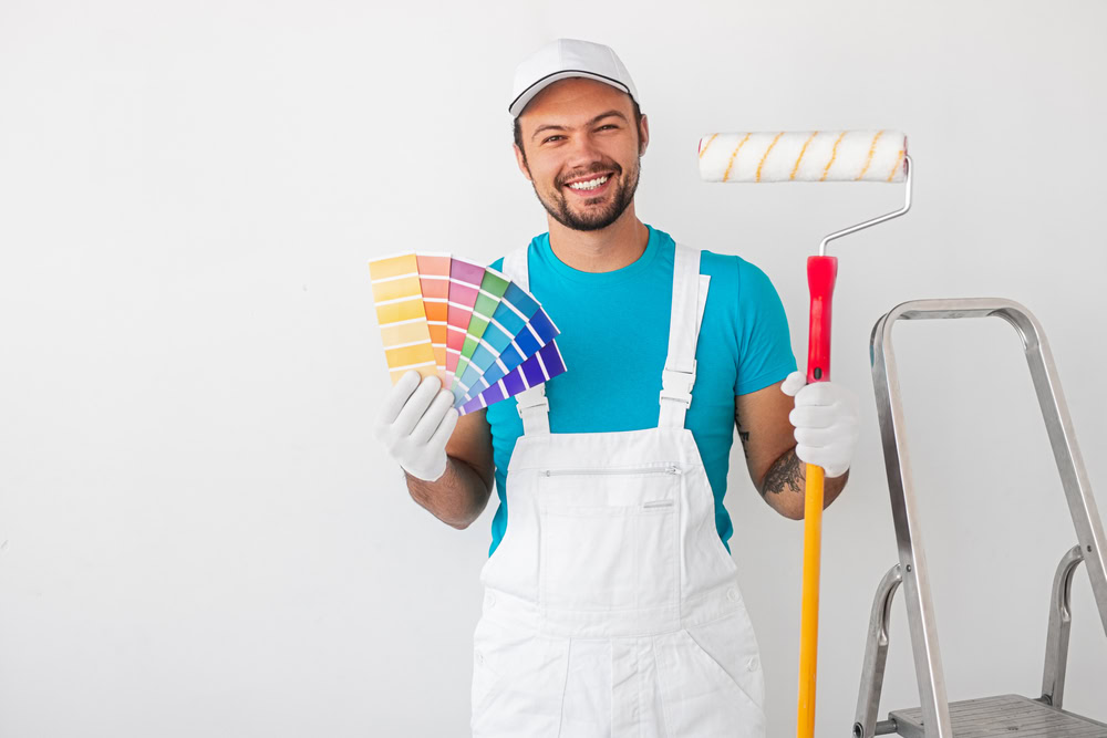 A smiling man in a white cap and overalls holds a paint roller in one hand and a fan of color swatches in the other. Standing beside a ladder, poised to start interior painting in Suffolk County, he is ready to transform the white wall behind him.