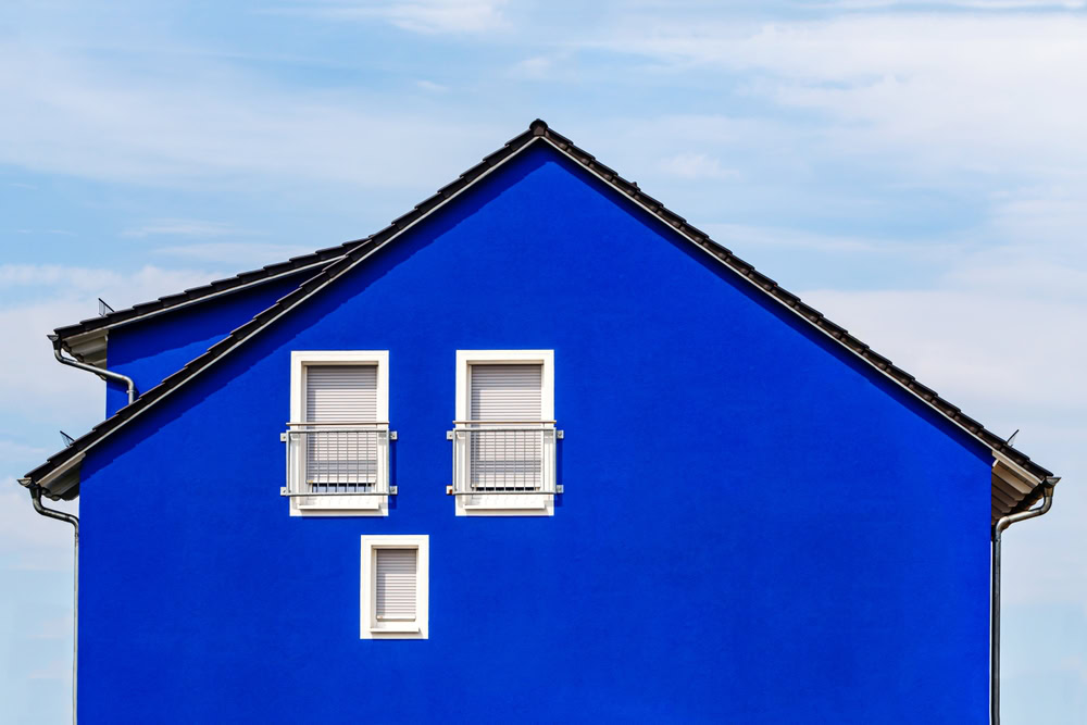 A bright blue house with a gabled roof stands proudly against a clear sky, showcasing its impeccable exterior painting typical of Nassau County. Two small white-framed windows with shutters and metal railings add charm to the side of the house.