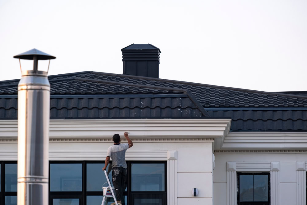 A person on a ladder is skillfully handling exterior painting in Suffolk County, transforming the building's facade beneath its dark tiled roof and large windows. A chimney and vent punctuate the roofline under a clear sky, showcasing meticulous artistry in every brushstroke.