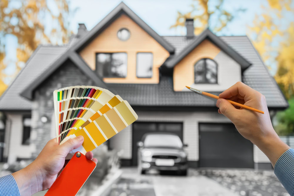 A person holds up a color palette and a paintbrush in front of a modern house, suggesting an exterior painting selection for renovation in Nassau County. The two-story house, with a black roof and contrasting exterior, stands elegantly amidst the vibrant autumn foliage.