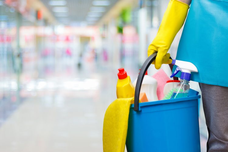 A person wearing yellow gloves is holding a blue bucket filled with various cleaning supplies, such as spray bottles and detergent. The background hints at recent interior painting in Suffolk County, with a brightly lit hallway slightly out of focus.